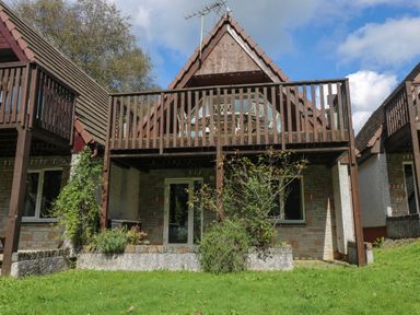 A two-story house with a wooden balcony a grassy lawn and trees at Devine Lodge in Callington