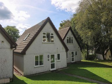 Two small houses with sloped roofs and white exterior walls on a grassy area with trees in the background at Devine Lodge in Callington