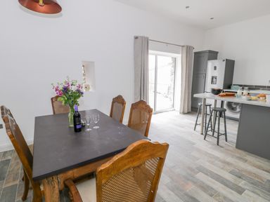 A dining area with a wooden table and chairs with flowers and wine on the table next to a kitchen with stools and appliances at Storws in Pentraeth