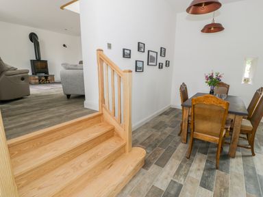 A dining area with wooden chairs and table with flowers next to wood stairs and a living room with sofas and a wood stove at Storws in Pentraeth