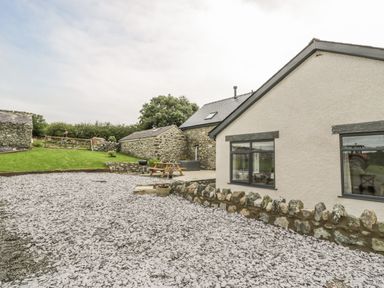 An outdoor area with a gravel driveway a stone wall a patio with a picnic table and a charcoal grill adjacent to a house and stone buildings at Storws in Pentraeth