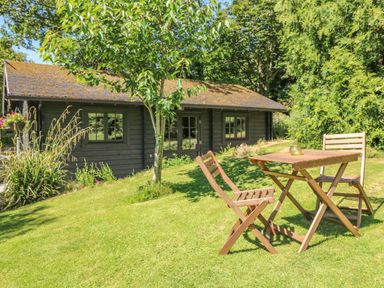 A wooden table and two chairs on grass with a tree and a cabin in the background at Pond Cabin in Stithians