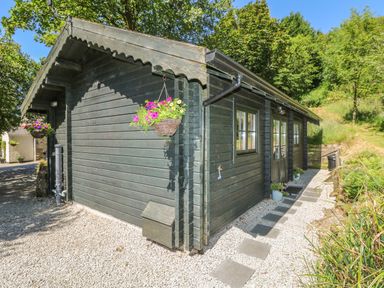 A wooden cabin with hanging flower baskets and a stone pathway surrounded by trees at Pond Cabin in Stithians