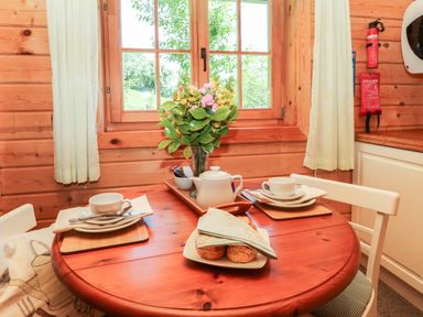 A wooden dining table with teacups plates a teapot and a plate of scones near a window with curtains at Pond Cabin in Stithians