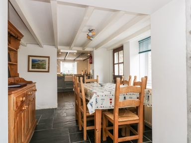 A dining area with a wooden table and chairs next to a window and a kitchen visible through an open doorway at Forge Cottage in Sennen