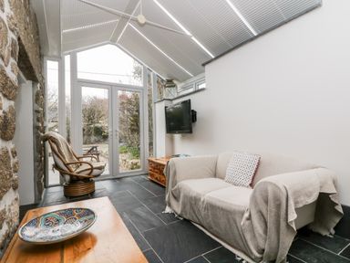 A room with a beige sofa covered with a throw and a cushion a rattan chair a wooden table with a decorative bowl and a wall mounted TV at Forge Cottage in Sennen