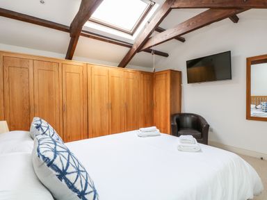 A bedroom with a bed a skylight wooden wardrobes a wall mounted television and a black chair at Forge Cottage in Sennen