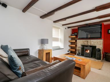 A living room with a leather sofa, wooden coffee table, side table with lamp, fireplace, wall-mounted TV, and window at Forge Cottage in Sennen