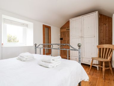 A bedroom with a metal bed frame folded towels on the bed a wooden wardrobe wooden chair and a window at Forge Cottage in Sennen