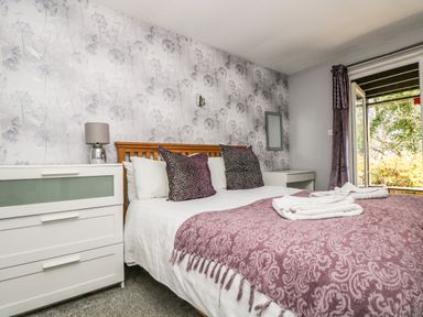 A bedroom with a double bed two pillows white and purple a bedside table a lamp a chest of drawers and an open door to a garden at Valley Lodge 58 in Callington