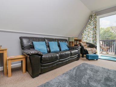 A living room with a black leather sofa with blue cushions a small black armchair with a brown throw a blue footstool wooden side tables a green lamp a bookshelf and a sliding glass door with floral curtains at Valley Lodge 5 in Callington
