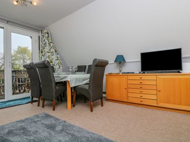 A dining area with a table set with wine glasses surrounded by dark chairs next to a wooden cabinet with a lamp and television at Valley Lodge 5 in Callington