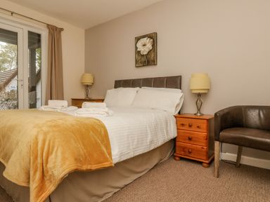 A bedroom with a bed with white sheets and yellow blanket bedside tables with lamps a leather chair and a door with curtains at Valley Lodge 5 in Callington