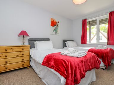 A bedroom with two single beds red blankets white linens a wooden dresser with a lamp and a window with red curtains at Valley Lodge 5 in Callington