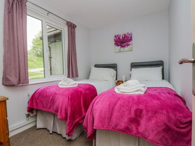 A bedroom with two single beds covered in pink blankets and white towels under a window with pink curtains at Valley Lodge 5 in Callington
