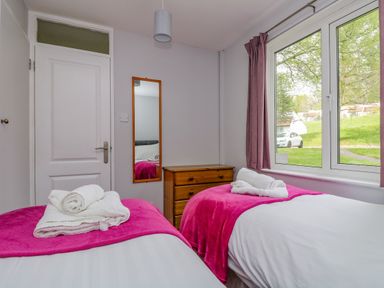 A bedroom with two single beds with pink blankets a wooden chest of drawers a mirror a door and a window with pink curtains at Valley Lodge 5 in Callington