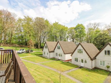 A balcony with wooden chairs overlooking a grassy area with pathways and white houses surrounded by trees at Valley Lodge 5 in Callington