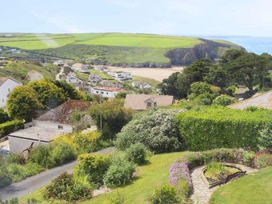 A coastal village with houses and a garden with a pond and flowers overlooking a beach and green hills at Windrush in Mawgan Porth
