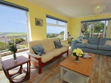 A living room with couches a coffee table with flowers and large windows showing an outdoor view at Windrush in Mawgan Porth