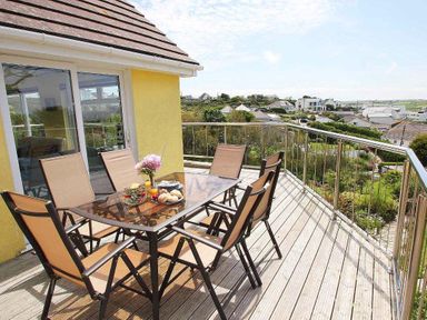 A balcony with glass table and six chairs overlooking a residential area at Windrush in Mawgan Porth