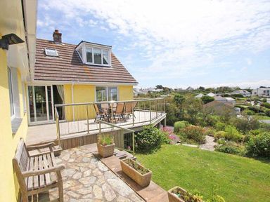 A yellow house with a balcony and outdoor chairs overlooking a garden and neighboring houses at Windrush in Mawgan Porth