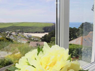 A coastal village with houses green fields and a sandy beach visible through a window with yellow flowers on the windowsill at Windrush in Mawgan Porth