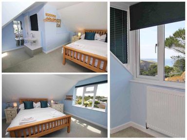 A bedroom with a wooden bed and bedside tables next to windows with views of trees and rooftops at Windrush in Mawgan Porth