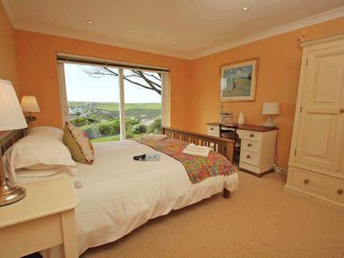 A bedroom with a bed a desk and chair a wardrobe and large window showing greenery at Windrush in Mawgan Porth