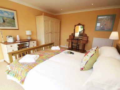 A bedroom with a wooden bed a colorful throw a wardrobe a desk with a lamp and a dressing table with a chair at Windrush in Mawgan Porth
