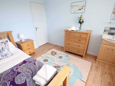 A bedroom with a wooden bed with patterned bedding a wooden dresser with a flower vase and a bedside table with a lamp at Windrush in Mawgan Porth