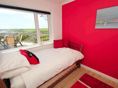 A bedroom with a single bed facing a large window showing an outdoor deck with chairs and a green landscape at Windrush in Mawgan Porth
