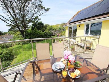 An outdoor patio with a glass table set for a meal and beige chairs next to a yellow house with solar panels at Windrush in Mawgan Porth