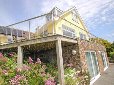 An exterior view of a yellow house with a stone lower level balcony and pink and white flowers outside at Windrush in Mawgan Porth