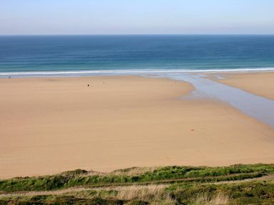 A sandy beach with a small stream flowing into the ocean and a grassy path in the foreground at Windrush in Mawgan Porth