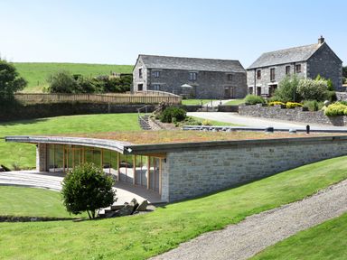 An outdoor view of a modern low building with glass walls and stone exterior surrounded by green grass and two stone houses in the background at Marhayes in Camelford