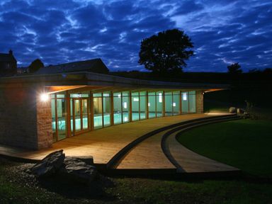 An illuminated indoor swimming pool with glass walls and wooden deck outside at Marhayes in Camelford