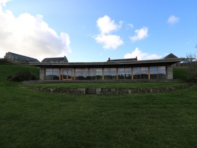 A single-story building with large glass windows on a grassy hill with stone houses in the background at Marhayes in Camelford