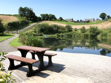 A picnic table on a wooden deck by a pond with grassy hills and houses in the background at Marhayes in Camelford