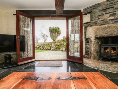 A room with a wooden door open to a garden a stone fireplace with a fire and a television on a stand at Westhayes in Camelford