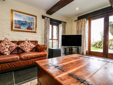 A living room with a brown leather sofa with patterned cushions a wooden chest coffee table a television on a stand floral curtains and wooden framed glass doors at Westhayes in Camelford