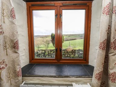 A window with wooden frame and black tiled sill showing a green landscape outside with stone wall and curtains with tree patterns at Westhayes in Camelford