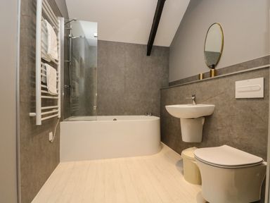 A bathroom with a bathtub and shower, towel rack with towels, wall-mounted sink, round mirror, toilet, and trash bin at Westhayes in Camelford