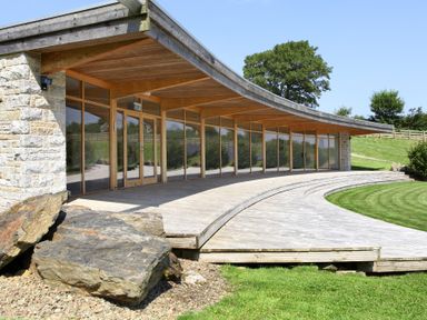 A curved wooden deck with large glass windows and stone walls outside a building with grass and trees around at Westhayes in Camelford