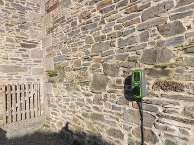 A stone wall with an electric vehicle charging station and a wooden gate behind gravel at Westhayes in Camelford
