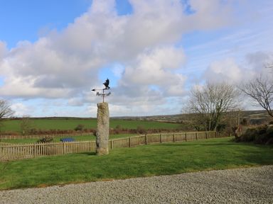 A garden with a stone pillar topped by a weather vane a wooden fence trees and green fields at Westhayes in Camelford