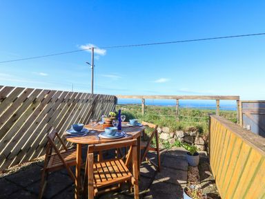 An outdoor patio with a wooden dining table and four chairs set with tableware overlooking a grassy area and ocean at Kerensa ha-Kres in Sennen