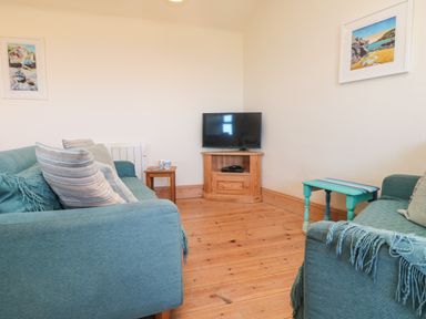 A living room with two blue sofas a wooden TV stand with a television a small wooden side table and paintings on the wall at Kerensa ha-Kres in Sennen
