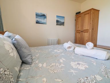 A bedroom with a bed covered in floral bedding with rolled towels on top two paintings on the wall a wooden wardrobe and a heater Kerensa ha-Kres Sennen