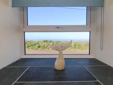 A window with a whale tail decoration on the windowsill overlooking greenery and the sea at Kerensa ha-Kres in Sennen