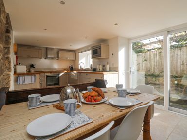 A dining table set with plates cups a teapot and croissants in a kitchen with cabinets and large glass doors at Bal View in St Just